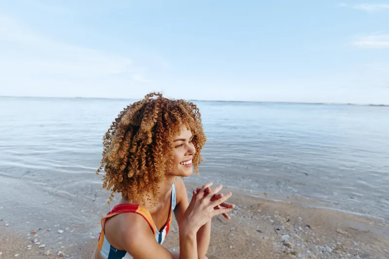 Beach ocean woman curly hair smile summer travel relaxation, on sandy shore by calm waves, she makes a gentle hand gesture, outdoor sunlight scene, peaceful happiness concept