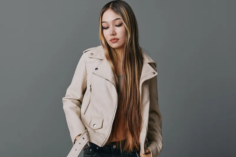 Young woman with long hair wearing light brown leather jacket and brown sweater standing against gray background with serious expression and casual style fashion portrait.