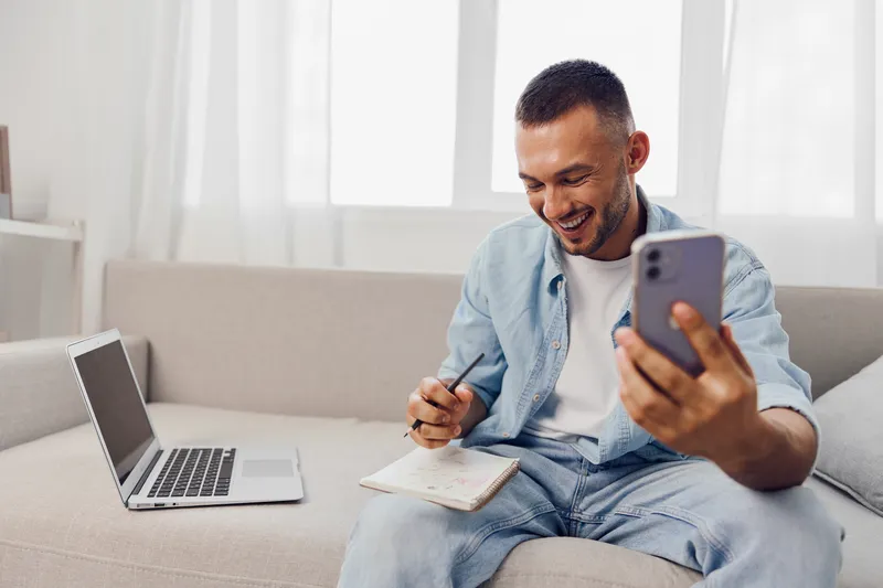 Smiling man engaged in a creative brainstorming session, jotting down ideas in a notebook while using his smartphone, capturing modern work dynamics and inspiration.