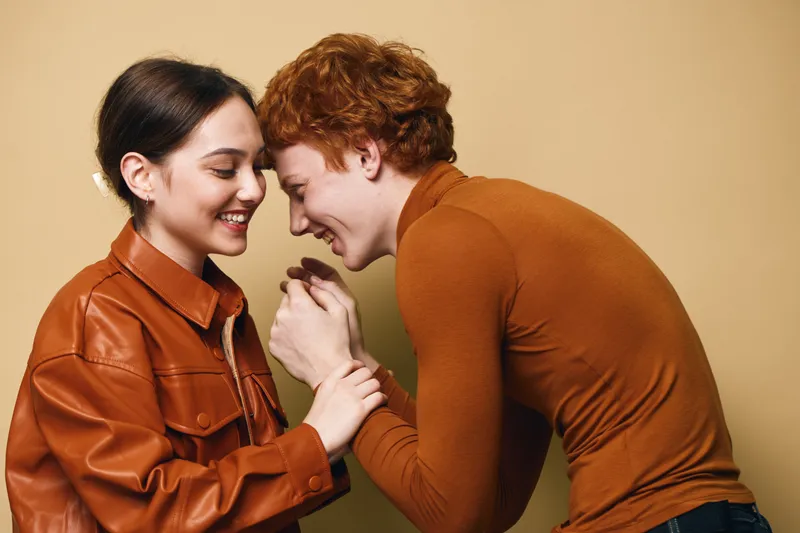 Young couple smiling and holding hands close together in warm autumn clothing standing against beige background. Intimate moment with happiness and affection between man and woman.