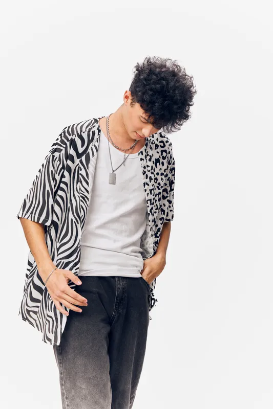 Young man with curly hair wearing a black and white animal print shirt and white t-shirt, standing with head down against a clean white background.