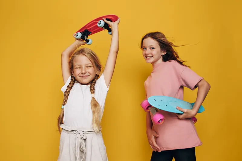Two happy girls with skateboards pose on a yellow background. They smile and show casual style with relaxed attitude.