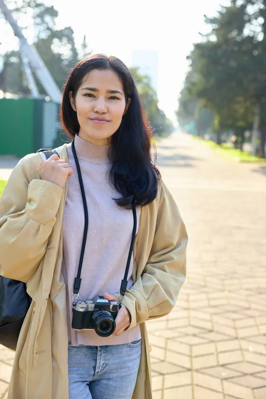 Camera in view as a woman stands outdoors on a sunny path, casual style, wearing a beige coat and knit top, ready for street photography and travel shoots, capturing candid daily life.