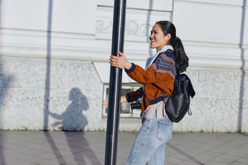 Woman with backpack holds a street pole in an urban outdoor setting, casual fashion, smiling expression, daylight, and everyday city life on a sunny day