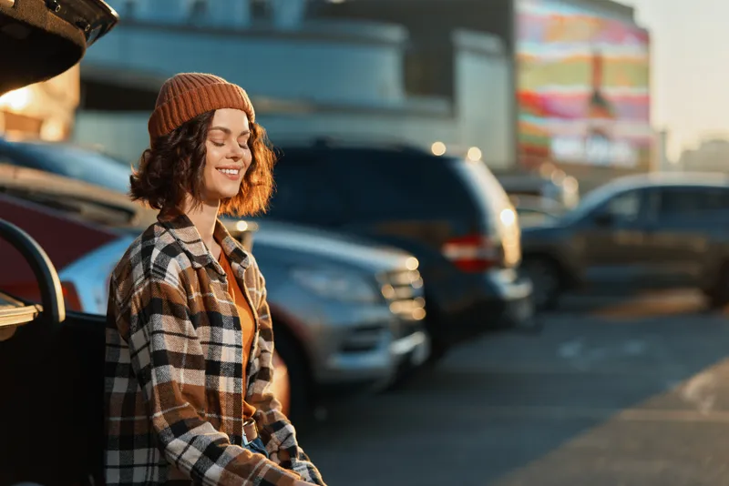 Woman portrait smile with eyes closed on car trunk in urban parking at golden hour, candid lifestyle and authenticity, mindful living and emotional storytelling with warm glow.