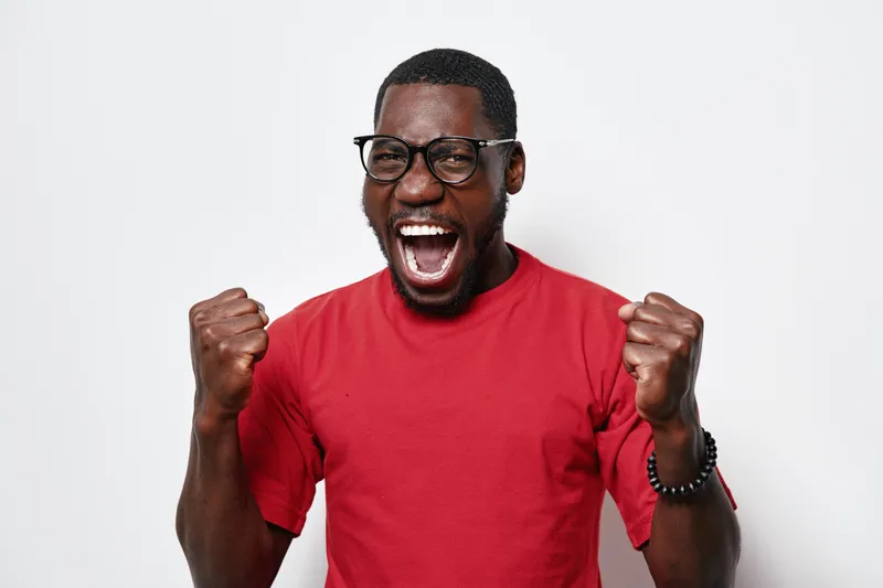African American man with dark skin wearing glasses and red t shirt celebrating success with clenched fists and open mouth on white background. Excited, happy and energetic expression.