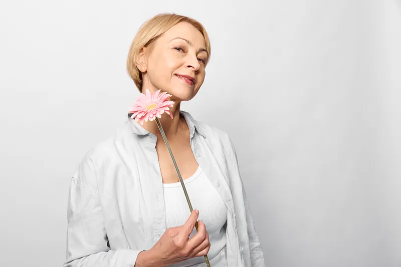 Elderly woman with pink flower, studio portrait closeup, smiling softly, white background, relaxed seated pose, spring mood, gentle light, calm expression