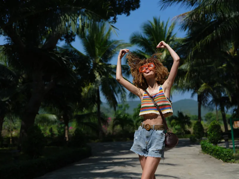 carefree young woman with curly hair wearing colorful top and denim shorts dancing outdoors, enjoying sunny day in tropical park with palm trees and clear blue sky