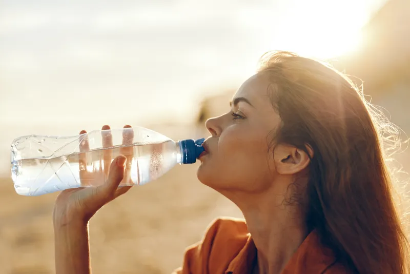 Woman drinking water bottle outdoors, sunlight and sun flare, summer hydration and health, calm profile portrait, wind in hair, warm desert sand background, wellness concept
