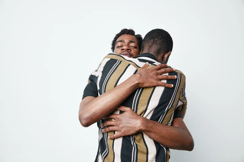 Two african men hugging each other with eyes closed and emotional connection, dark skin friends showing support and friendship in casual striped shirts on white background.