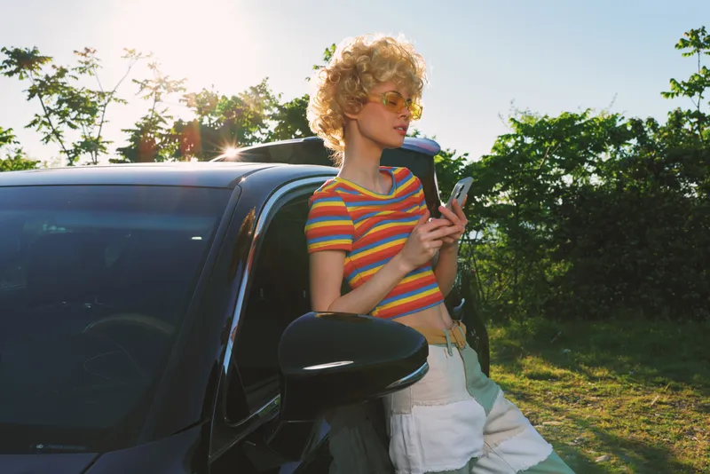A woman leans against a dark car in a sunny outdoor setting, wearing a striped tee and sunglasses. A retro color palette enhances a casual lifestyle moment.