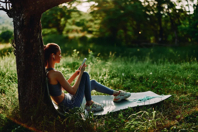 Young woman in activewear enjoying a smoothie while relaxing outdoors on a yoga mat in a sunlit park, emphasizing healthy lifestyle and mindfulness