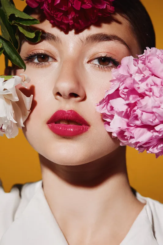 Close-up portrait of a young woman with bold red lips and flawless skin surrounded by colorful peony flowers on a mustard background. Beauty, fashion, and creative makeup concept.