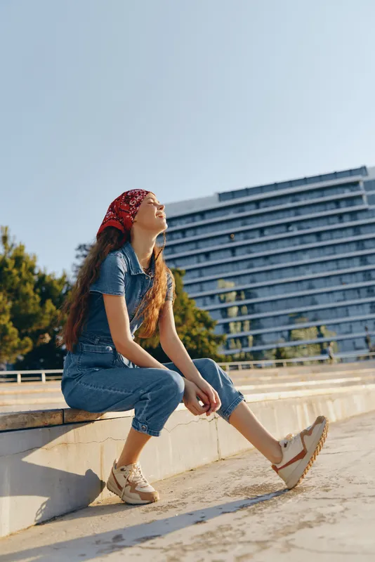 Lifestyle woman in modern boho-western denim street style sitting outdoors on a sunny day with film color tones, wearing casual sneakers and a red bandana.