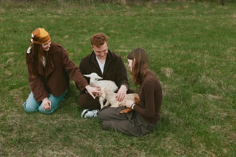 Friendly trio sits on grass outdoors, guiding a young goat kid as they connect with smiles, casual fashion, warm daylight, relaxed lifestyle, intimate moment in a rustic park