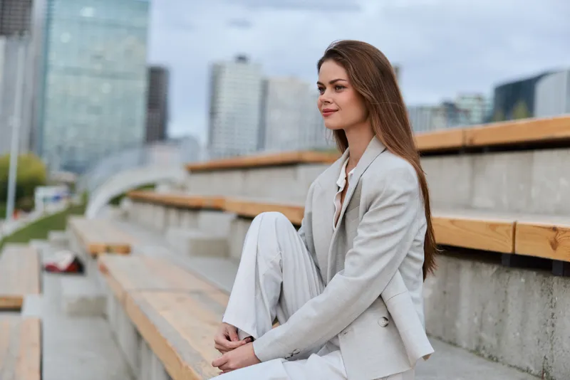 Confident woman in business attire sitting outdoors with a city skyline background, showcasing professionalism and poise in a dynamic urban setting.