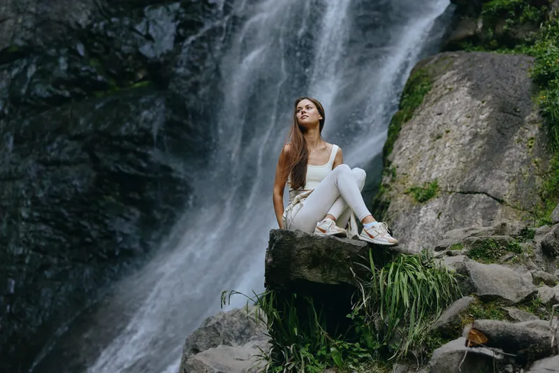 woman sitting waterfall outdoors, dressed in white casual clothes, surrounded by rocks and greenery, enjoying nature and peaceful moment in forest landscape.