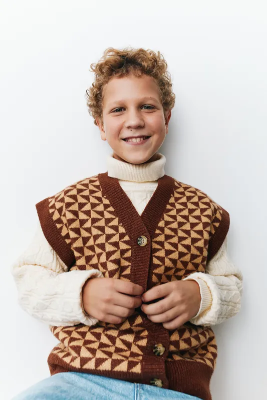 Authentic lifestyle portrait of a smiling child in a geometric knit vest, warm tones, casual school look, capturing a candid moment of youthful energy and charm.