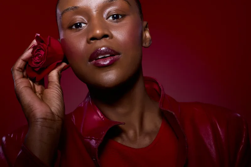 Close-up of a dark skinned African woman holding a red rose near her face against a red background. Concept of beauty, elegance, and natural skincare with vibrant colors and style.