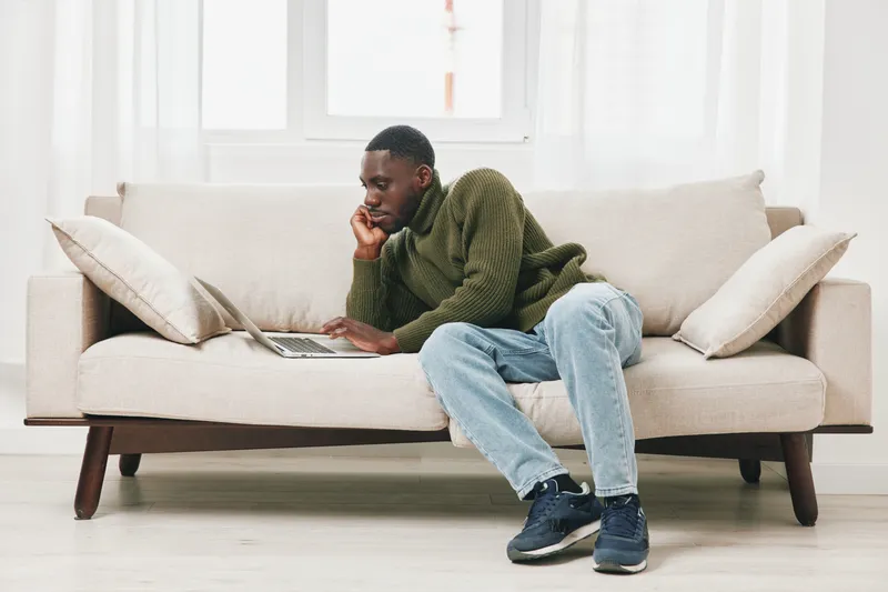 Thoughtful young man in a green sweater and jeans working on a laptop at home, showcasing a casual and focused atmosphere