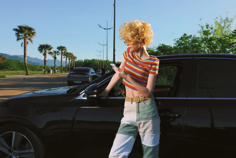 A young woman with curly blonde hair in a striped crop top and faded denim jeans leans on a sleek car in a sunny street, conveying retro chic and everyday confidence.