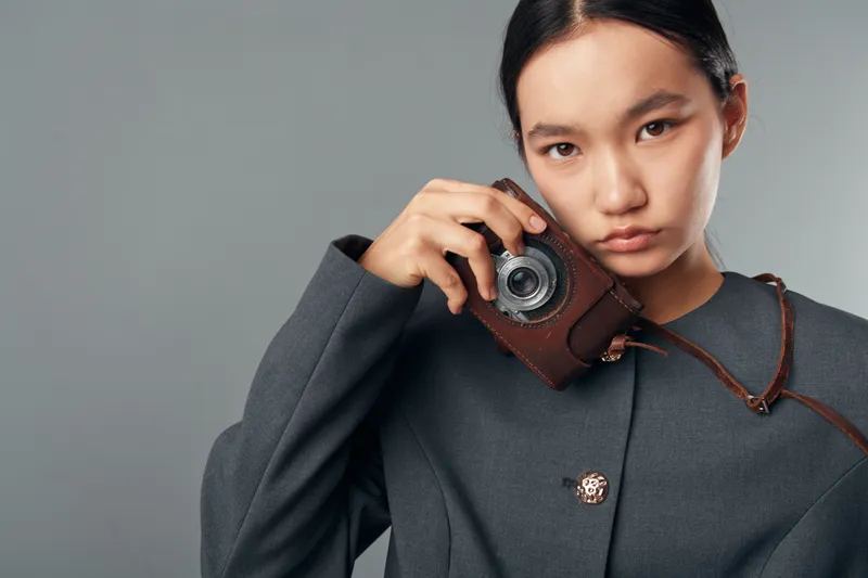Woman holding vintage camera close to face, wearing dark coat, posing with serious expression on gray background. Portrait shot showcasing retro photography equipment and fashion style.
