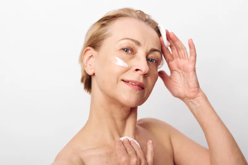 Mature woman with short blonde hair applying white face cream on cheek, smiling gently. Close-up on neutral background. Concept of skincare, aging beauty, self-care and wellness routine.