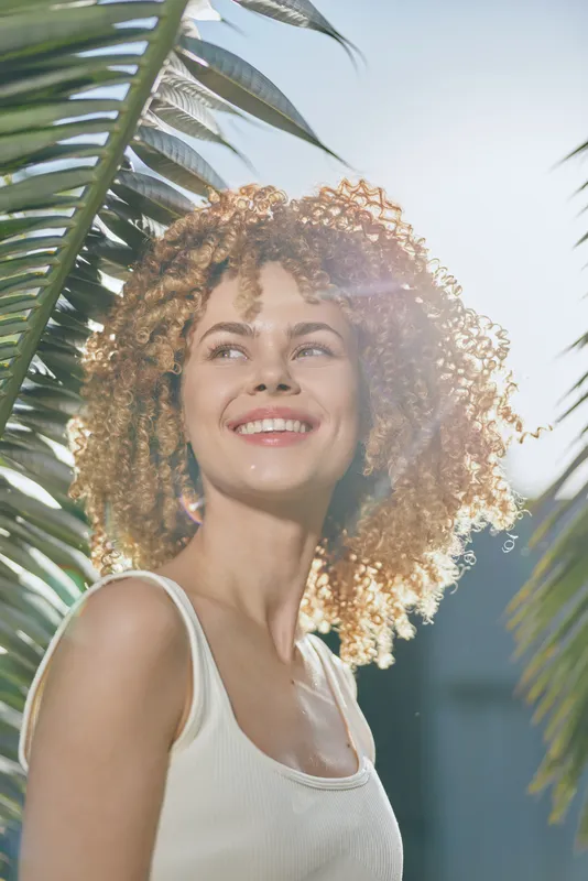 smiling woman with curly hair outdoors surrounded by green palm leaves, wearing white tank top, bright natural sunlight enhancing a cheerful summer vibe