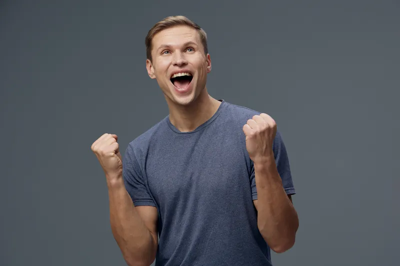 Enthusiastic young man celebrating success, expressing joy and excitement, wearing casual gray t-shirt, fists clenched, smiling widely, isolated on a plain gray background for lifestyle and happiness 