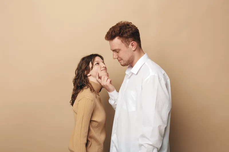 Young couple in casual clothes looks at each other with affection, man gently touches woman’s chin. Beige background creates warm and intimate atmosphere of love and connection.