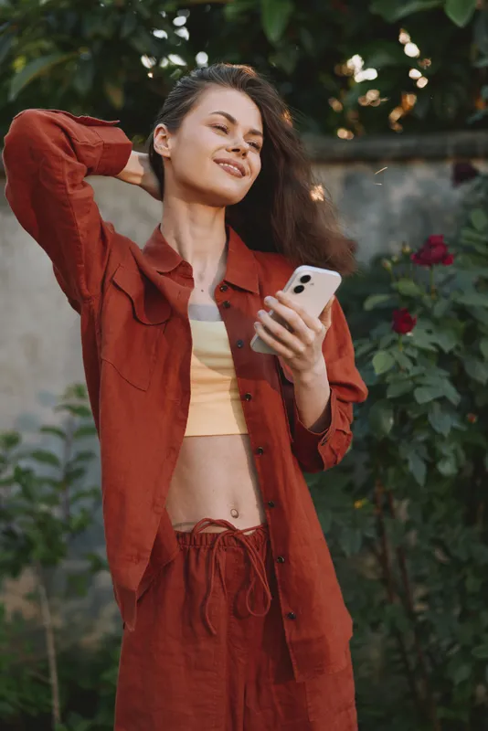 Smiling woman in rust-colored outfit holding a smartphone in a sunny garden, showcasing a relaxed lifestyle, casual fashion, and self confidence with warm natural light, outdoor leisure, modern