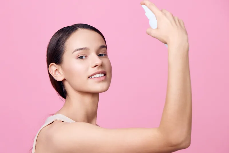 Young woman applying skincare product in a light, natural way against a pink background, showcasing a fresh and radiant appearance