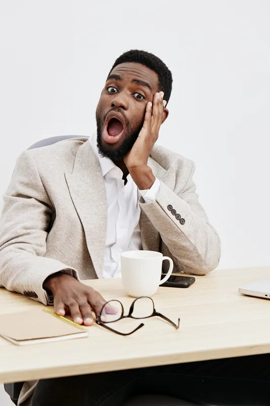Shocked young African American man in formal jacket with surprised facial expression, hand on cheek, sitting at desk with glasses, coffee mug, notebook, and smartphone, indoor studio portrait. People 