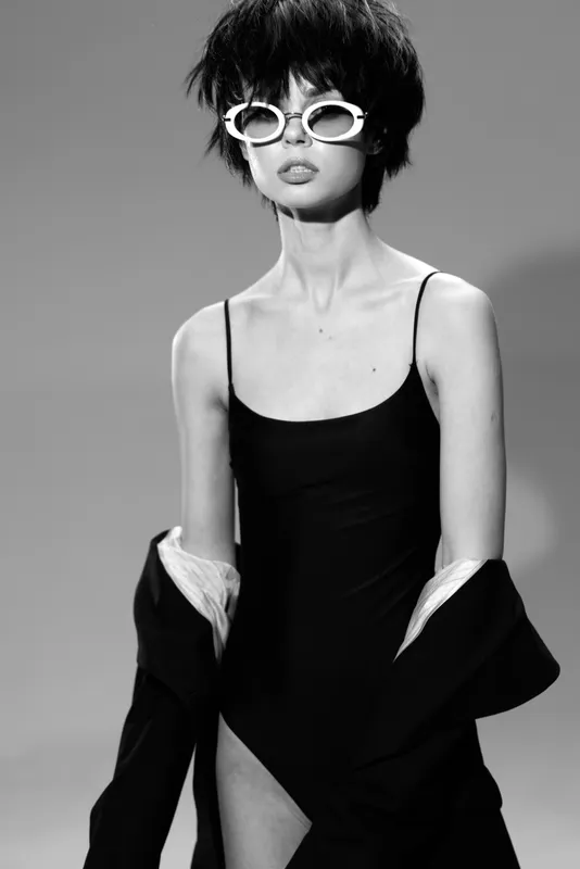 Black and white portrait of a stylish woman wearing white sunglasses and a black dress with a jacket, posing confidently in a studio setting.