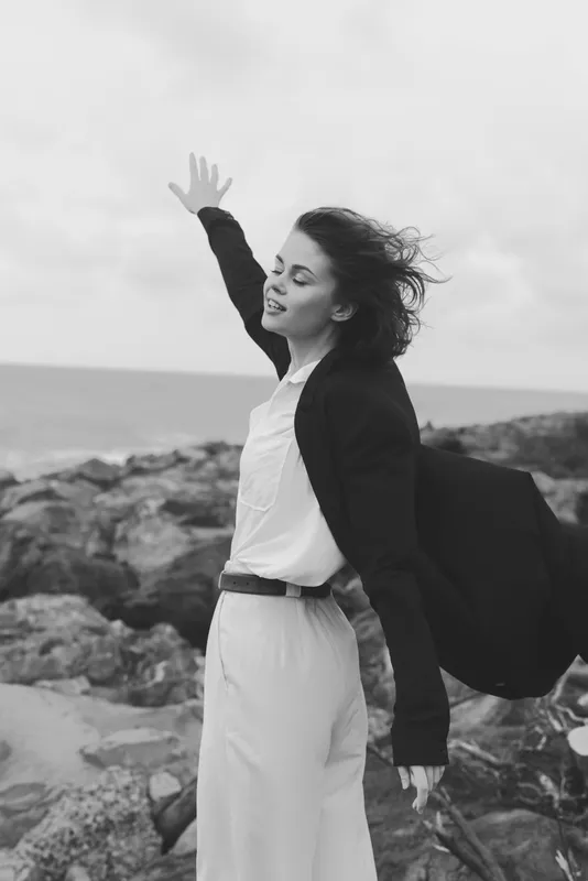 Woman on a windy coastal scene raises her hand, sea and rocks in the background, wearing a blazer over a light dress, expressing confidence and freedom in an outdoor moment