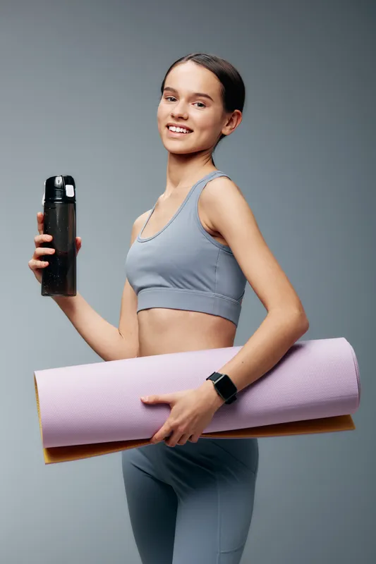 Fitness woman in sporty attire holding a water bottle and yoga mat, ready for training. Clean studio background highlights health, wellness and an active lifestyle.