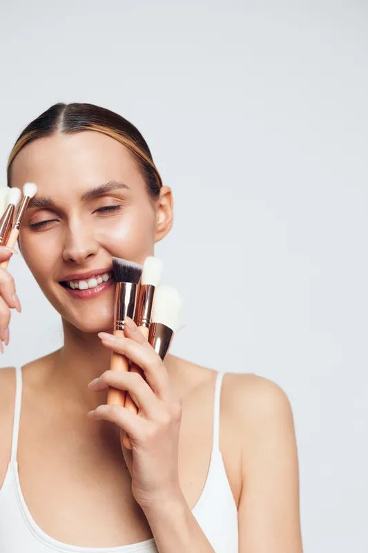 A smiling young woman with clear skin holds various makeup brushes near her face against a plain background. Concept for beauty, skincare, and makeup routine.