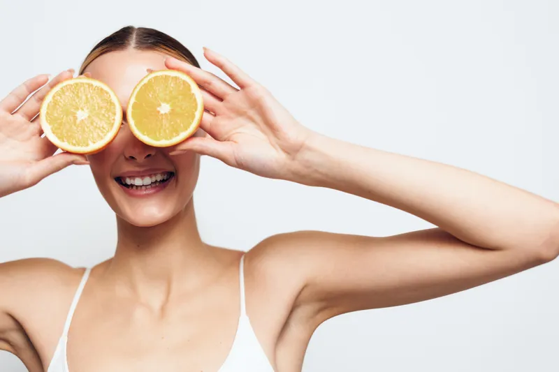 Young woman with brunette hair holds two halves of an orange in front of her eyes while smiling against a white background in a playful and fresh studio portrait.