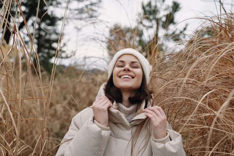 Woman in a warm beige coat and knit hat stands among tall grasses, smiling under a cloudy sky, capturing a cozy outdoor moment in late autumn or early winter soft breeze.