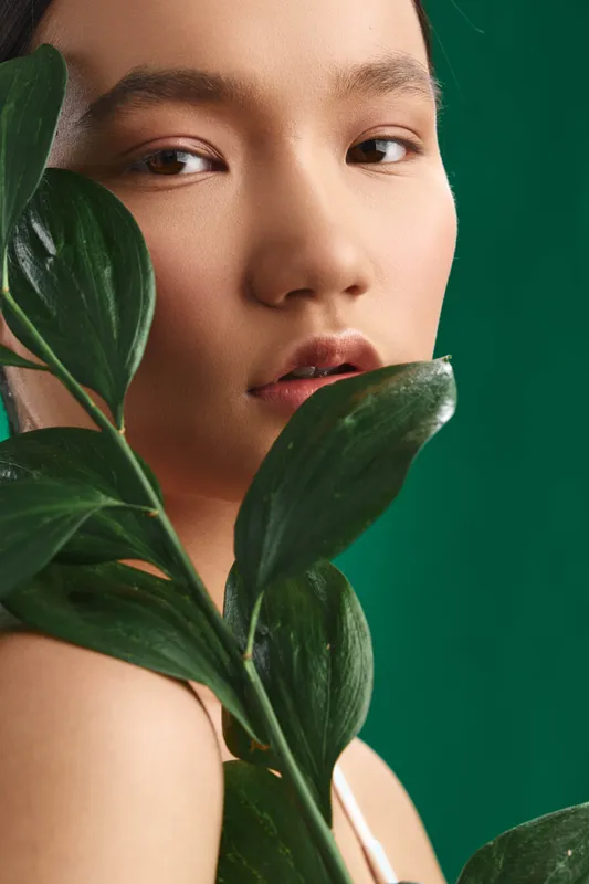 asian woman gazes softly through lush leaves against a deep green backdrop, conveying calm beauty and thoughtful presence in a studio portrait with natural light and serene mood