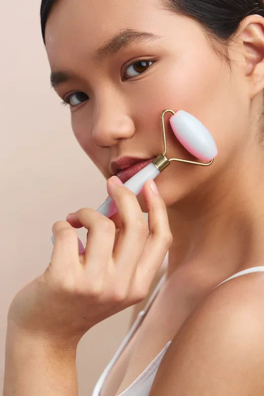 asian woman applies a facial roller during a calm skincare session, showcasing a focused, serene expression in a bright, minimalist space with soft lighting.