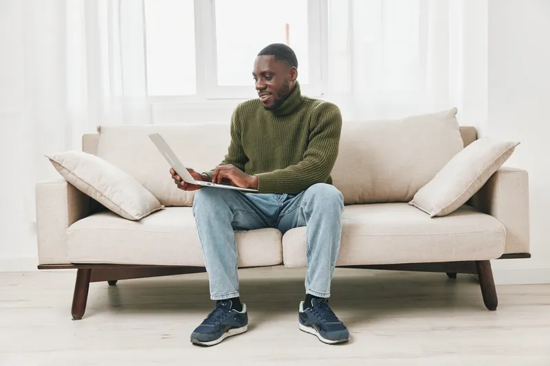 Young African man working on a laptop at home, dressed in a green sweater, sitting on a light sofa, conveying a relaxed and focused emotion while surrounded by a bright, cozy atmosphere
