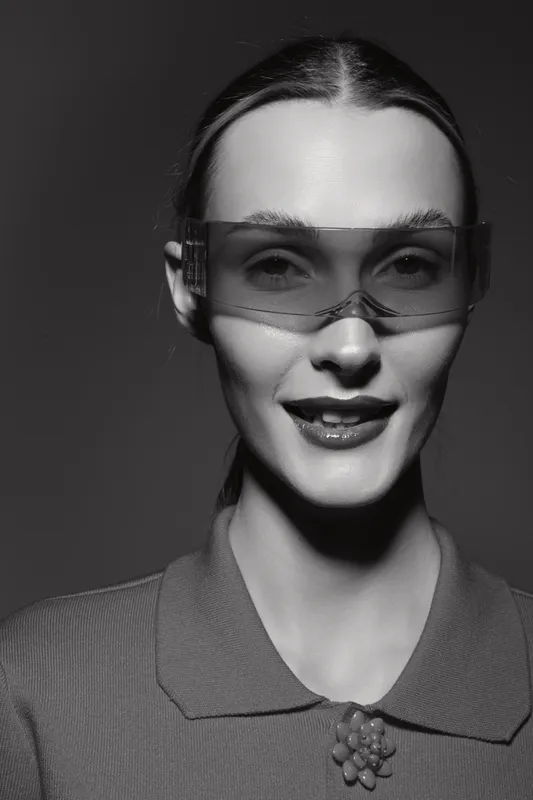 White woman with pale skin wearing futuristic sunglasses and a collared shirt smiles in monochrome studio portrait.