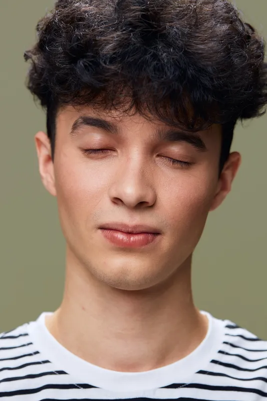 young man with closed eyes wearing striped shirt, calm expression, soft lighting, beige background, natural skin tone, short curly hair