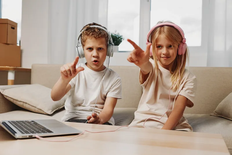 Two children wearing headphones enjoy music and point fingers while sitting on a couch in a cozy living room.