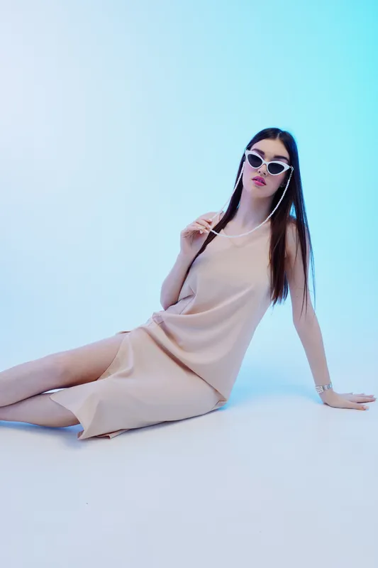 Young woman in beige dress and stylish sunglasses poses sitting on the floor in a bright studio. Concept of woman beauty fashion and modern style with soft lighting.