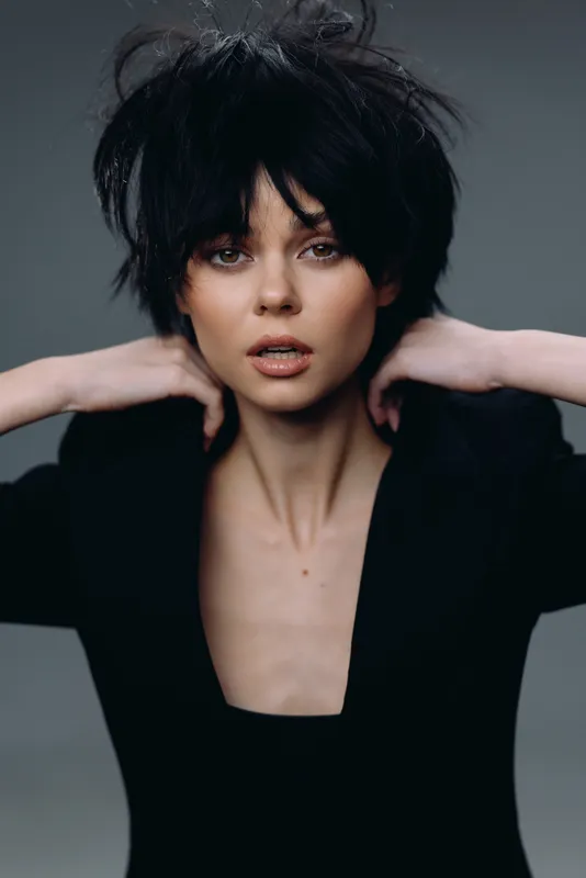 Young woman with short black hair posing confidently in dark clothing, studio portrait with soft lighting.