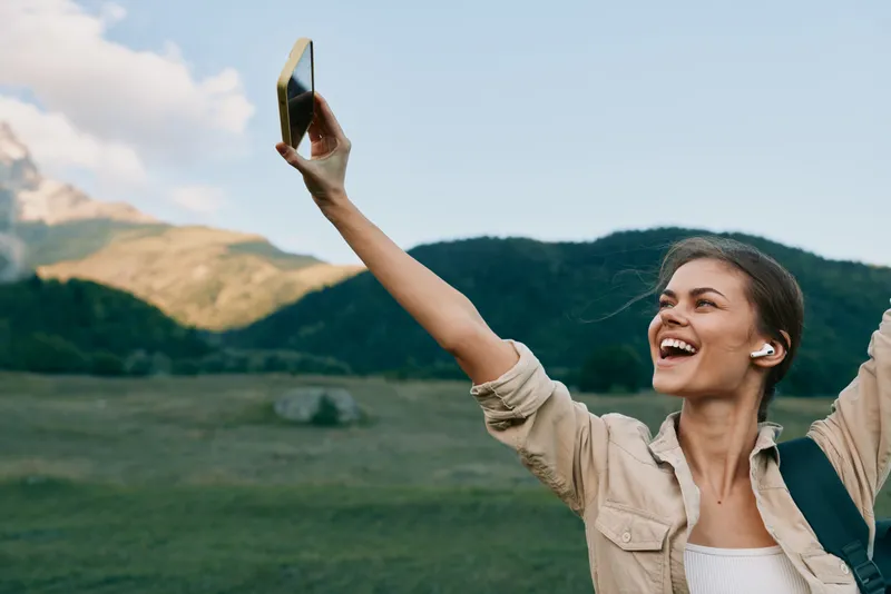 A happy young woman in casual clothes takes a selfie outdoors with mountains in the background. She wears wireless earbuds and enjoys a sunny day in nature.