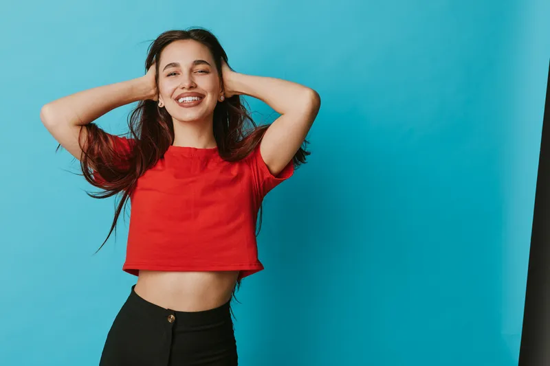 Young woman smiling with closed eyes and hands behind head in red crop top and black pants against blue background for happiness and casual style portrait.