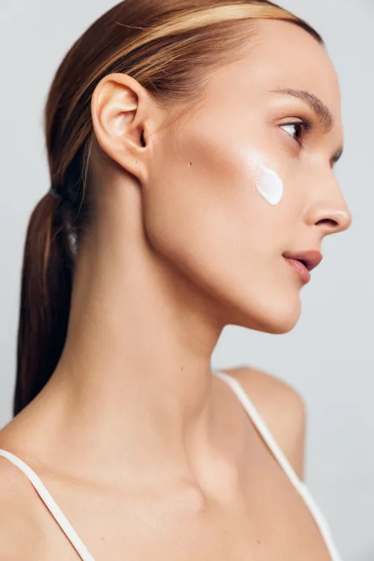 Young woman with clear skin applies white cream on cheek closeup natural beauty skincare routine isolated on white background minimal makeup and healthy complexion